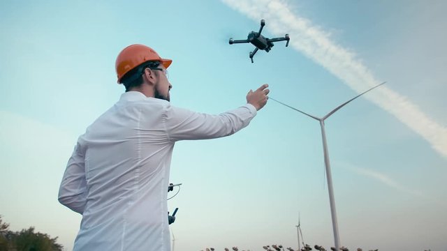 Caucasian engineer with a beard in an orange protective helmet and goggles landing smart quadrocopter drone on the background of an electric power wind generator in a green field. Slow Motion.