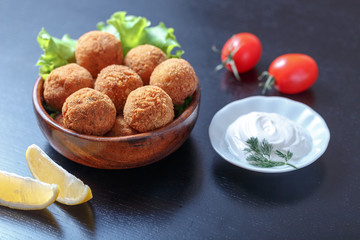 Falafel lies in a wooden plate. On the table lie tomatoes, cucumbers, lettuce, dill, lemon, sour cream.