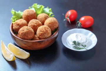 Falafel lies in a wooden plate. On the table lie tomatoes, cucumbers, lettuce, dill, lemon, sour cream.