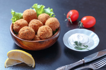 Falafel lies in a wooden plate. On the table lie tomatoes, cucumbers, lettuce, dill, lemon, sour cream.