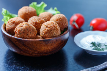 Falafel lies in a wooden plate. On the table lie tomatoes, cucumbers, lettuce, dill, lemon, sour cream.