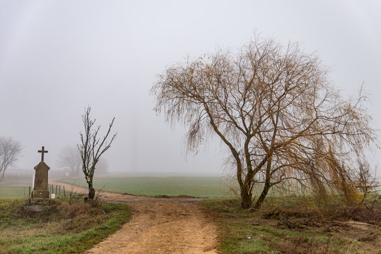 Petite stèle avec une croix et un arbre dans la brume au bord d'une route de Moselle