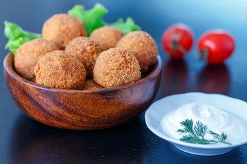 Falafel lies in a wooden plate. On the table lie tomatoes, cucumbers, lettuce, dill, lemon, sour cream.