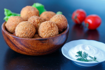 Falafel lies in a wooden plate. On the table lie tomatoes, cucumbers, lettuce, dill, lemon, sour cream.
