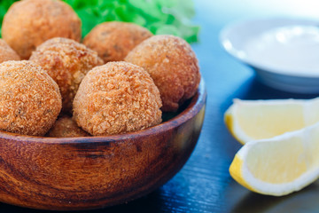 Falafel lies in a wooden plate. On the table lie tomatoes, cucumbers, lettuce, dill, lemon, sour cream.
