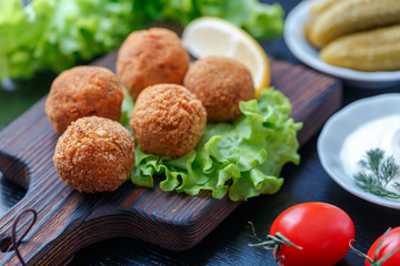 Falafel lies on a wooden cutting board. On the table lie tomatoes, cucumbers, lettuce, dill, lemon, sour cream.