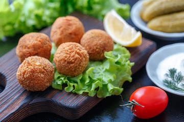 Falafel lies on a wooden cutting board. On the table lie tomatoes, cucumbers, lettuce, dill, lemon, sour cream.