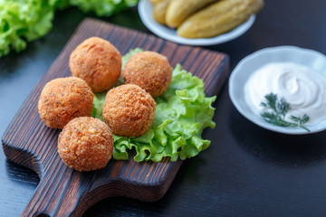 Falafel lies on a wooden cutting board. On the table lie tomatoes, cucumbers, lettuce, dill, lemon, sour cream.