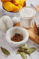 Spices in marble mortar and heap of bay leaves, jar with sea salt on cutting board, bowl with lemons on marble surface