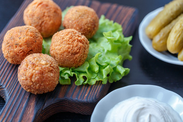 Falafel lies on a wooden cutting board. On the table lie tomatoes, cucumbers, lettuce, salad, dill, lemon, sour cream.