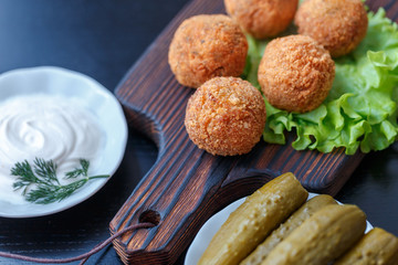 Falafel lies on a wooden cutting board. On the table lie tomatoes, cucumbers, lettuce, salad, dill, lemon, sour cream.