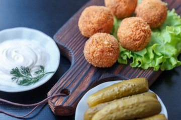 Falafel lies on a wooden cutting board. On the table lie tomatoes, cucumbers, lettuce, salad, dill, lemon, sour cream.