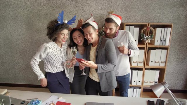 Multiethnic group of young women and men taking selfie on smartphone and smiling happily while celebrating Christmas in office
