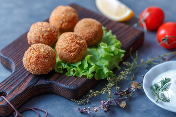 Falafel lies on a wooden cutting board. On the table lie tomatoes, cucumbers, lettuce, salad, dill, lemon, sour cream.