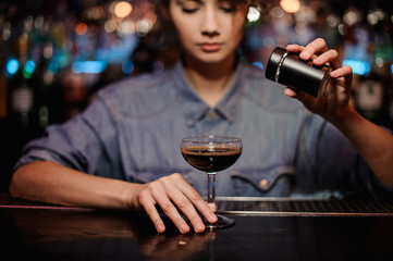 Female bartender adding to a cocktail with brown alcohol a powder on the bar counter