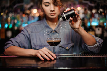 Bartender girl adding to a cocktail with brown alcohol a powder on the bar counter