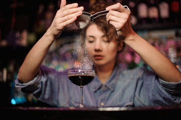 Bartender girl adding to a cocktail with brown alcohol a powder through the strainer on the bar counter
