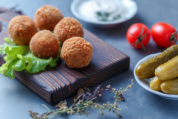 Falafel lies on a wooden cutting board. On the table lie tomatoes, cucumbers, lettuce, salad, dill, lemon, sour cream.