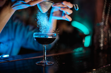 Bartender girl adding to a cocktail decorated with badian a sugar powder through the strainer in the blue light