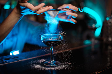 Female bartender adding to a cocktail decorated with badian a sugar powder through the strainer in...