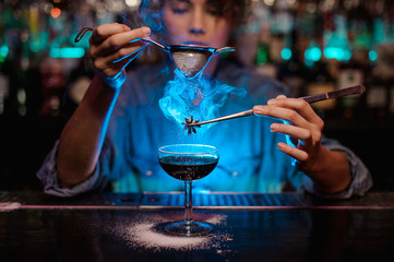 Girl adding to a brown cocktail and pour on a flamed badian on tweezers a powdered sugar through the strainer in the blue light