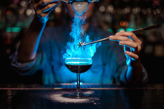 Bartender girl adding to a brown cocktail and pour on a flamed badian on tweezers a powdered sugar in the blue light