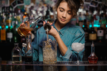 Female bartender pouring to the measuring glass cup with ice cubes an alcoholic drink from steel jigger