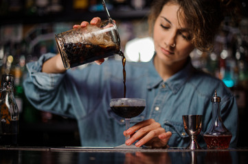 Girl pouring a brown alcoholic cocktail from the measuring cup to the glass