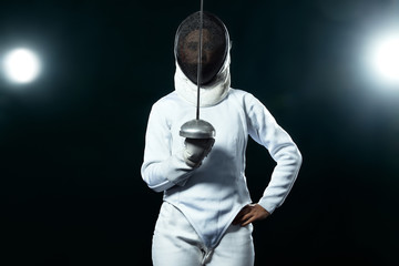 Young fencer athlete wearing fencing costume holding the sword and mask. Isolated on black background with lights - obrazy, fototapety, plakaty