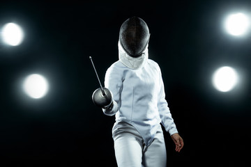Young fencer athlete wearing fencing costume holding the sword and mask. Isolated on black...
