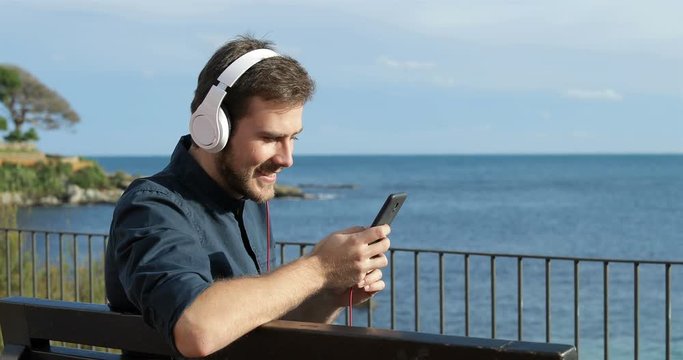 Happy man wearing headphones listening to music from a smart phone sitting on a bench on the beach