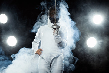 Young fencer athlete wearing fencing costume holding the sword and mask. Isolated on black...