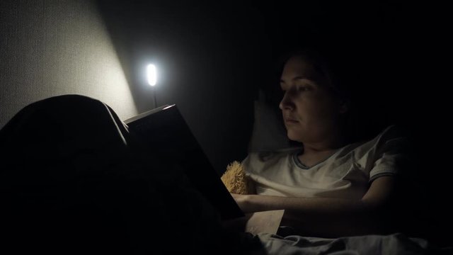 Beautiful caucasian girl reading book in bed at night with lamp light.