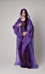 Naklejka premium full length portrait of beautiful girl with long black hair, wearing purple fantasy medieval gown and cloak. standing pose on grey studio background.