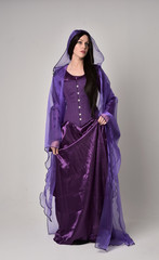 full length portrait of beautiful girl with long black hair,   wearing purple fantasy medieval gown and cloak. standing pose on grey studio background.