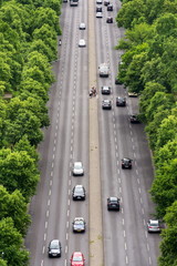 Cyclist waiting in the middle of busy road with cars passing from Brandenburg Gate on several lanes, aerial view with green trees on sides, Tiergarten, Berlin, Germany
