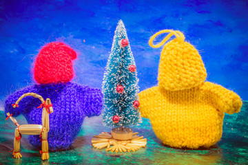 Two little man knitted by hand on the needles, yellow and blue are standing near a dressed-up Christmas tree.