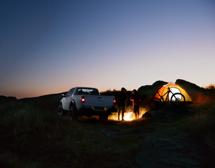 Friends near Bonfire, Pickup Truck, Tent and Bike at Night Camp in the Mountains. Adventure and Travel Concept