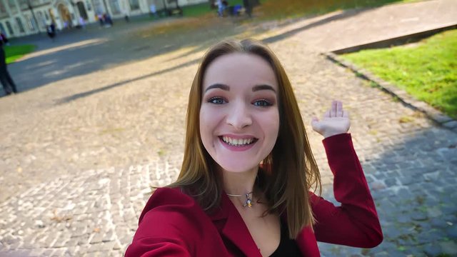 Young gorgeous girl student having a video call, showing space around her, waving with a hand, and being amused outside the university