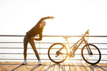 Photo of healthy woman doing stretching exercises, while standing near bicycle outdoors during...