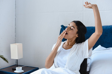 beautiful mixed race girl in white robe yawning and covering mouth in morning in bedroom - Powered by Adobe