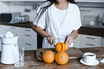 cropped image of mixed race girl in white robe cutting ripe oranges in morning in kitchen - Powered by Adobe