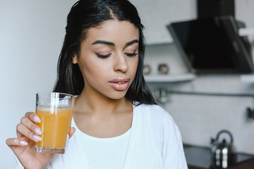 beautiful mixed race girl in white robe holding glass of fresh orange juice in morning in kitchen