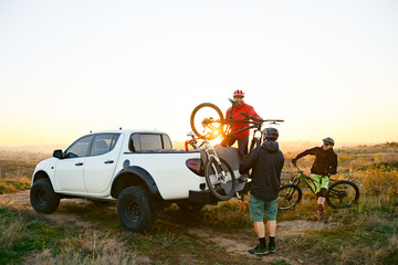 Friends Taking MTB Bikes off the Pickup Offroad Truck in Mountains at Sunset. Adventure and Travel Concept. - obrazy, fototapety, plakaty