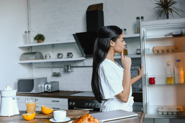 side view of pensive beautiful mixed race girl in white robe looking into fridge in morning in kitchen - Powered by Adobe