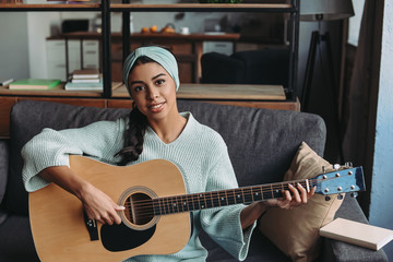 attractive mixed race girl in turquoise sweater and headband playing acoustic guitar on sofa at home and looking at camera - Powered by Adobe