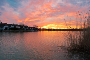 Naklejka premium Beautiful sunset at Lake Neusiedlersee in Austria with colorful orange and red illuminated cloudy sky. Picturesque holiday homes in terraced settlement. Reed grows on the shore.