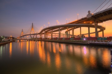 Naklejka na ściany i meble Low angle view of Bhumibol Suspension Bridge (or Industrial Ring Road ) over Chao Phraya River under evening twilight in Bangkok Thailand, with beautiful reflections on smooth water at red rosy dusk