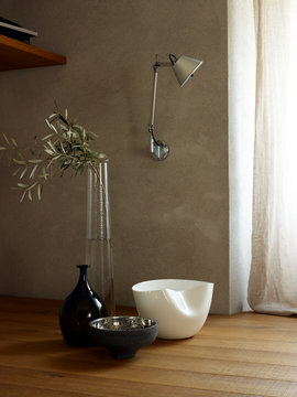 Olivenzweig in Glas Vase mit gefr&auml;stem Muster und Silber Schale und Porzellan Schale vor Wand mit Wandlampe und Fenster