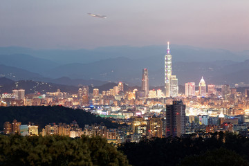Night scenery of Taipei, the vibrant capital city of Taiwan, with landmark  Tower standing amid skyscrapers, an airplane taking off from Songshan Airport & mountain silhouettes in evening twilight